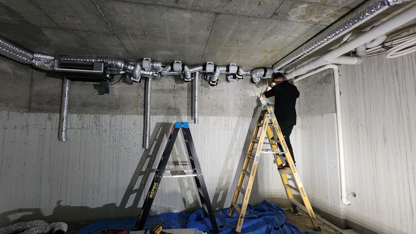 A long duct run going in across a basement ceiling, with ladders below