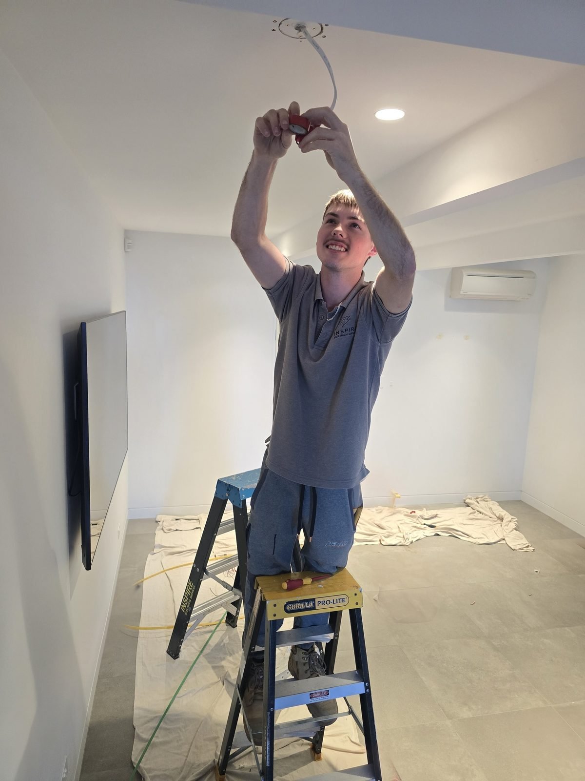 An electrician on a step ladder wiring a ceiling fitting, drop sheets on the floor